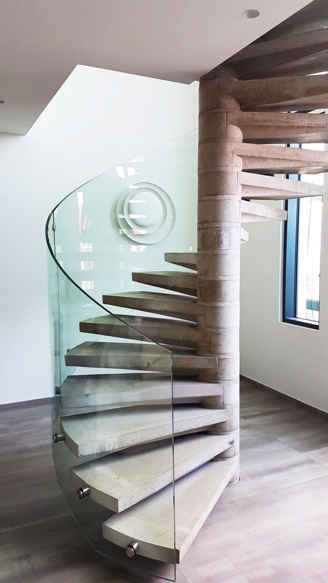 Escalier en colimaçon avec marches en bois, rampe en verre et colonne centrale texturée dans une maison moderne.