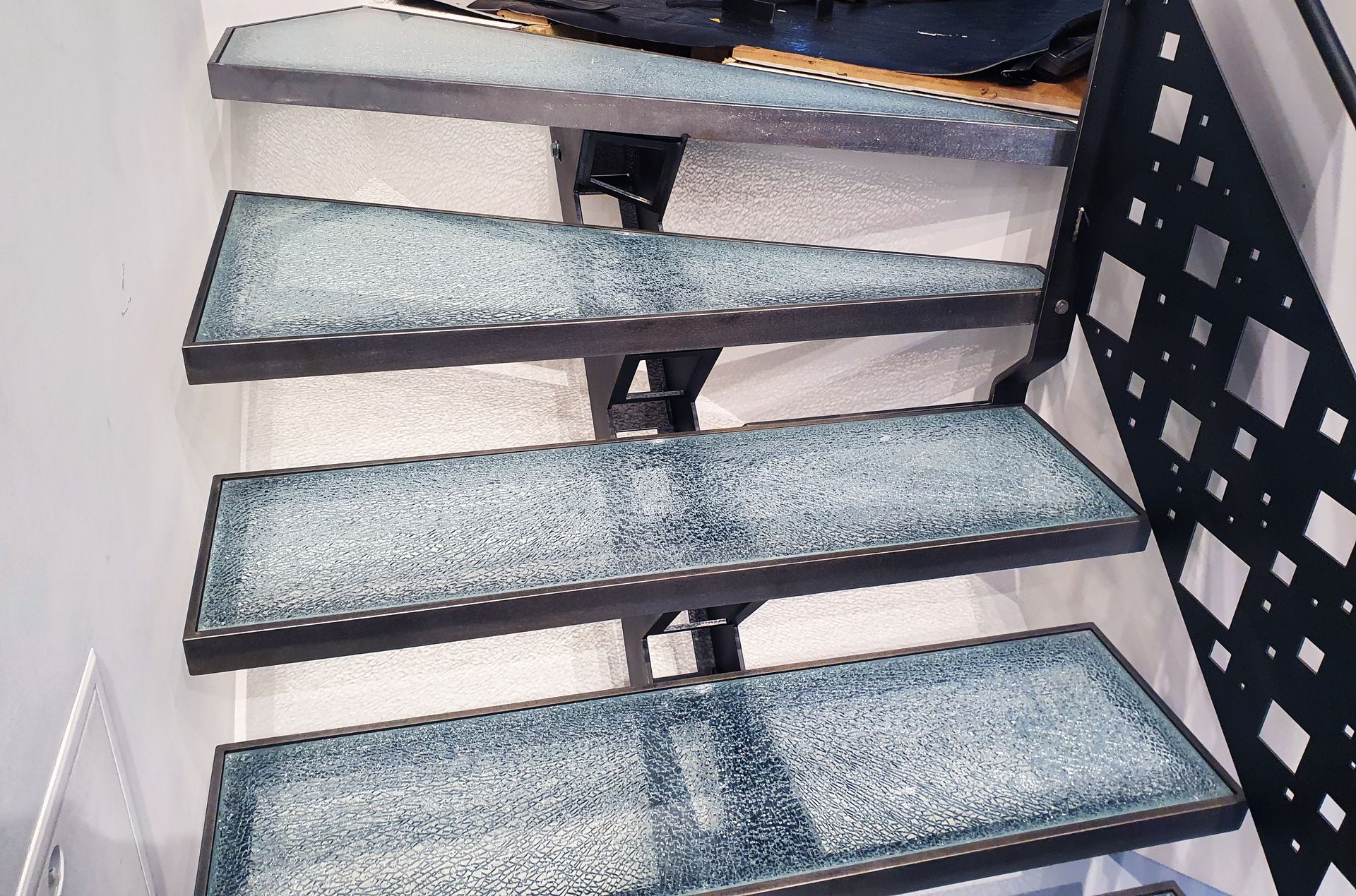 Escalier avec marches en verre et supports en métal noir, près d'une rampe géométrique noire, dans une pièce bien éclairée.