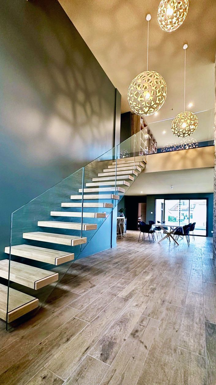 Escalier moderne avec rampe en verre, marches en bois, globes lumineux suspendus et mur bleu.