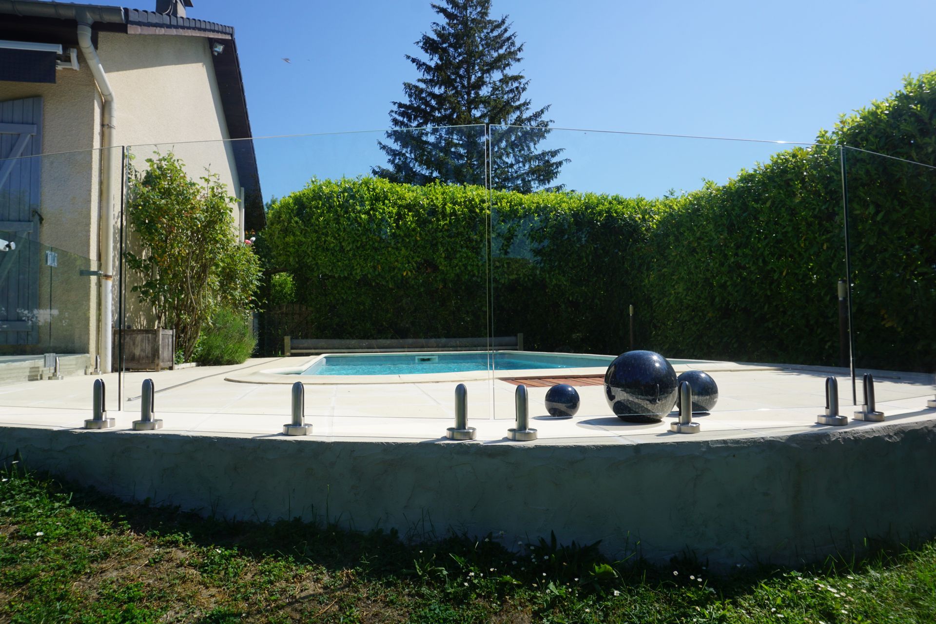 Piscine avec clôture en verre, entourée d'un patio en béton, d'une haie verte et d'une pelouse.