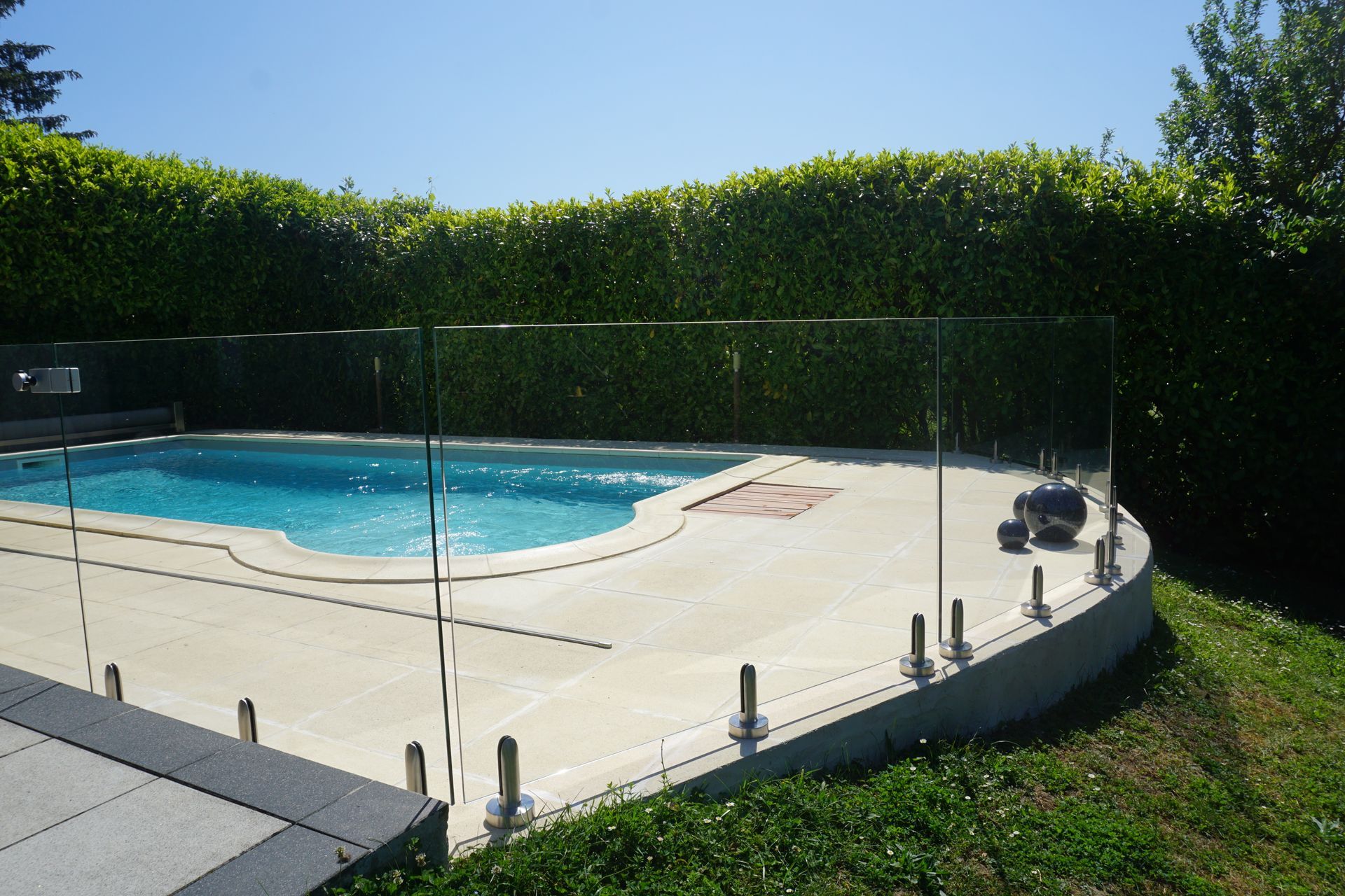 Une piscine entourée d'une clôture de sécurité en verre, avec une haie verte en arrière-plan.