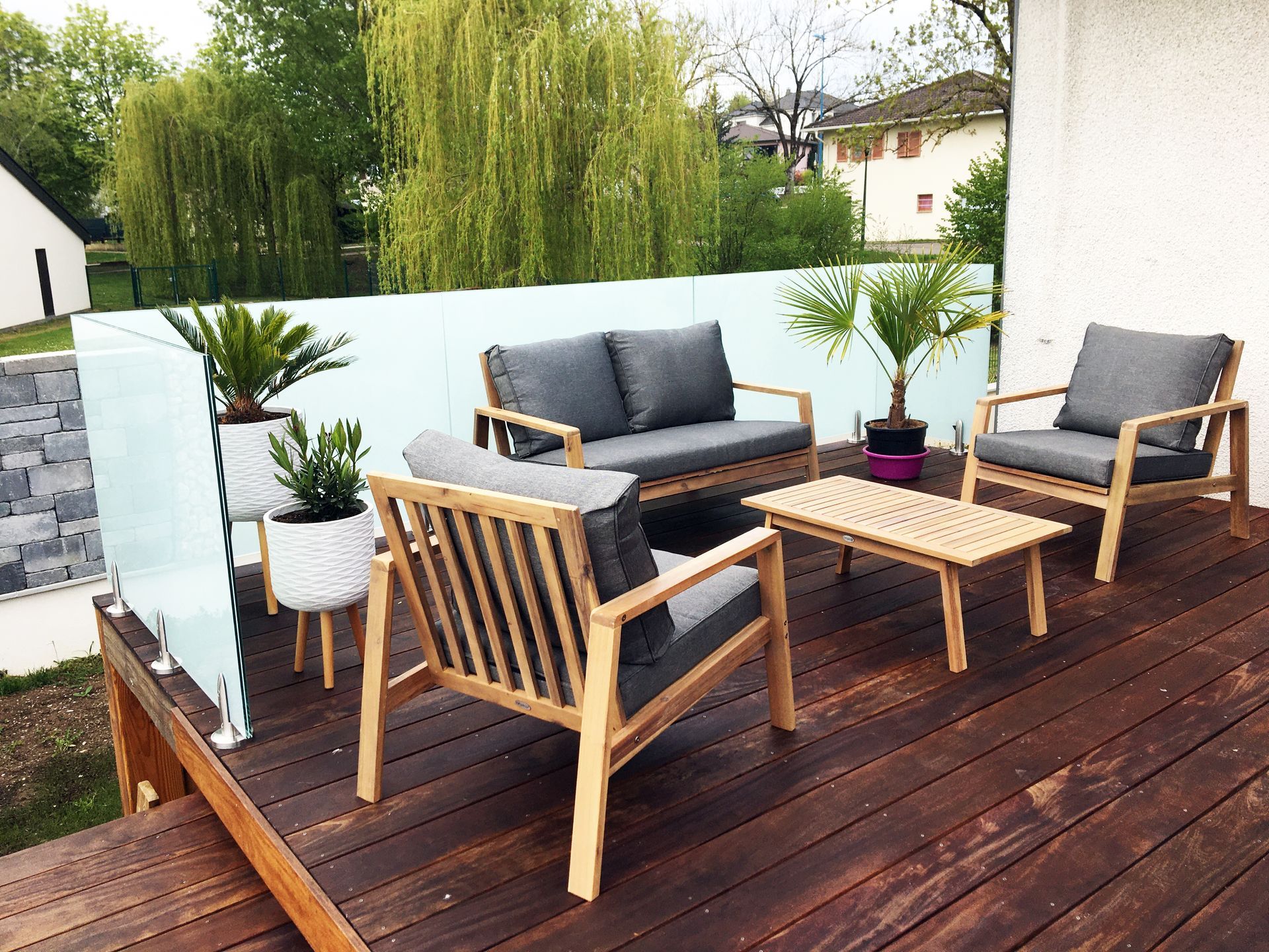 Terrasse extérieure avec mobilier en bois et plantes, donnant sur une cour arrière avec arbres et maisons.