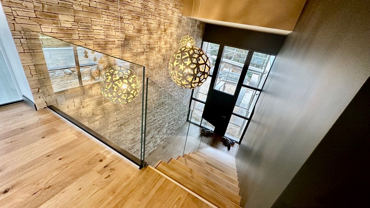 Escalier en bois avec rampe en verre, mur en pierre et lumières suspendues décoratives.