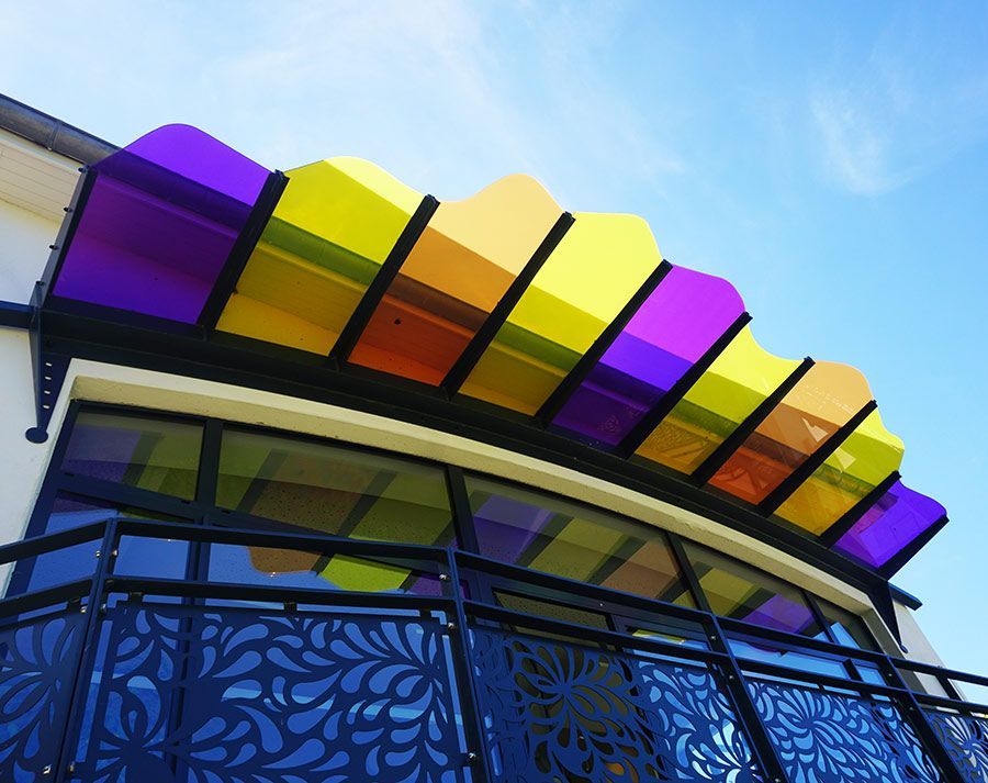Auvent coloré au-dessus de l'entrée d'un immeuble, avec des panneaux violets, jaunes et orange. Ciel bleu en arrière-plan.