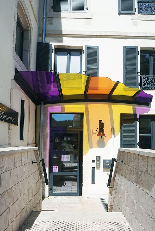Entrée avec auvent coloré, porte vitrée, marches en pierre et bâtiment blanc.