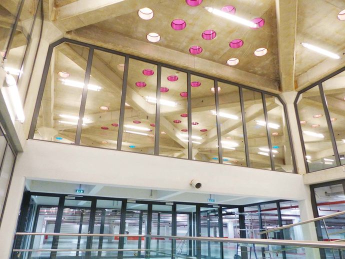 Intérieur de bâtiment moderne avec murs en verre, plafond en béton, lumières circulaires roses.