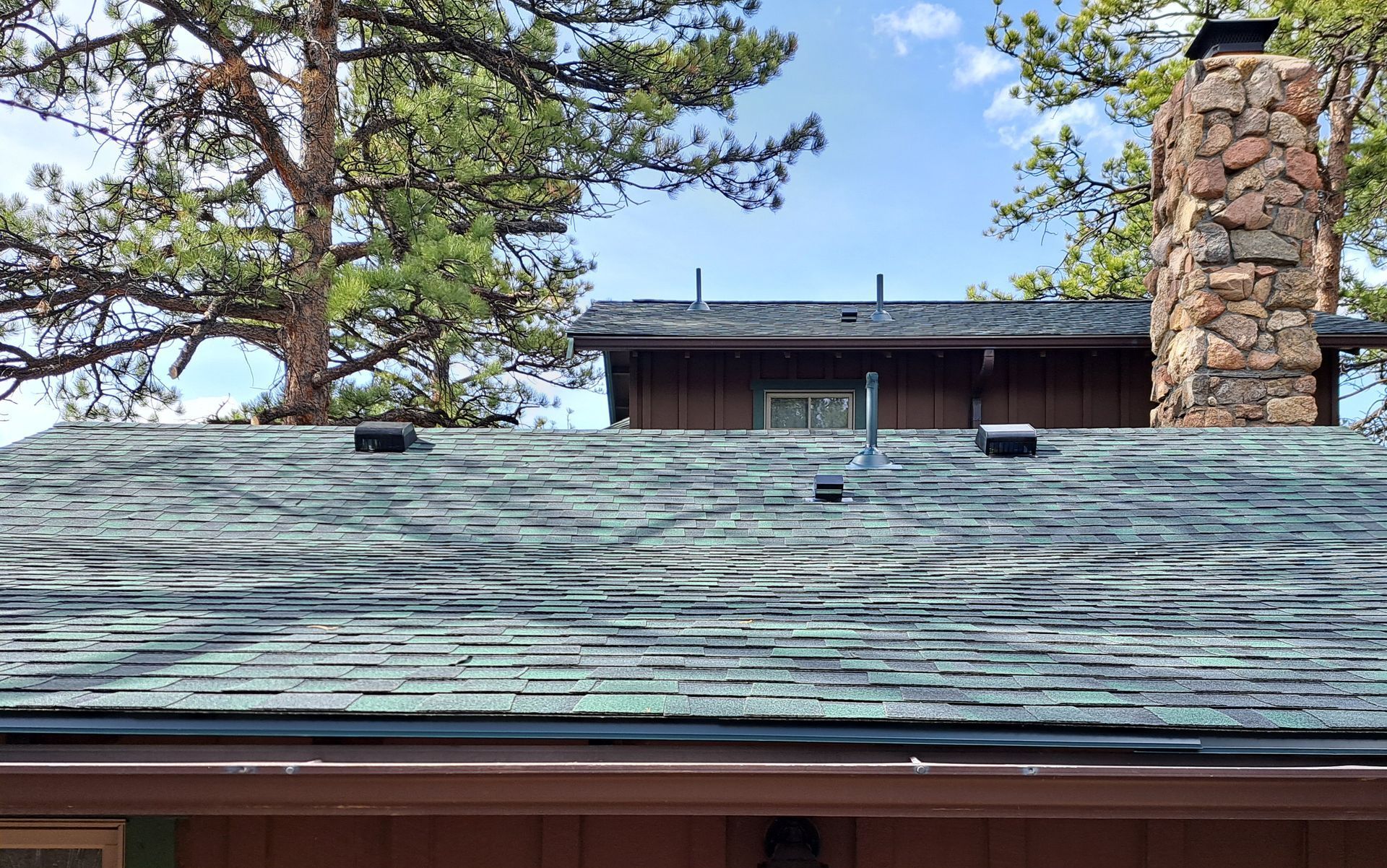 The roof of a house with a chimney and trees in the background.