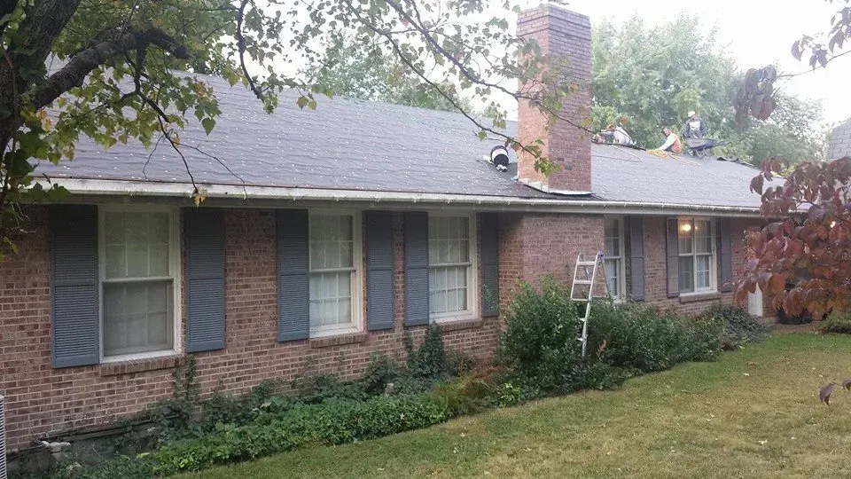 Brick house with blue shutters, front windows, chimney, and a sloped yard with trees around it