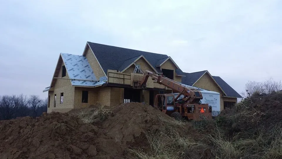 House under construction on a hillside, with exposed roof framing and a backhoe nearby