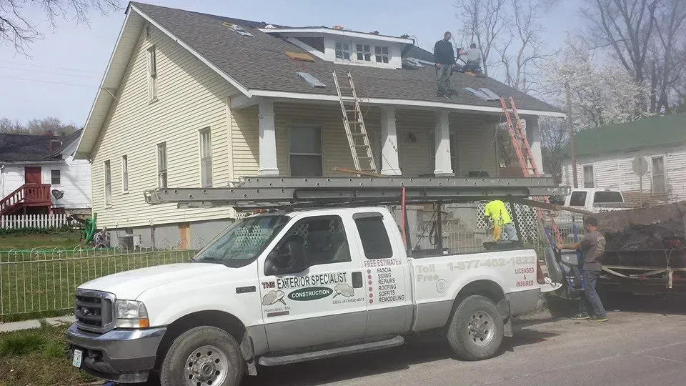 Utility truck parked beside a yellow house with workers on the roof carrying materials