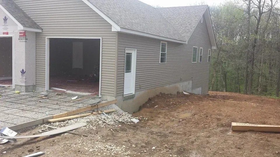 House exterior under construction with open garage, dirt yard, and wooded background