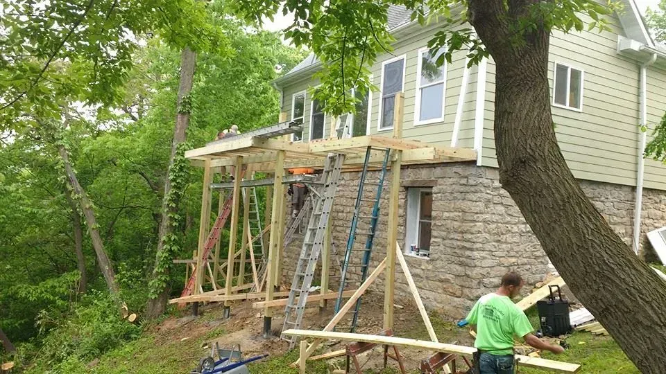 Workers build wooden supports under a house deck beside a green wooded yard.