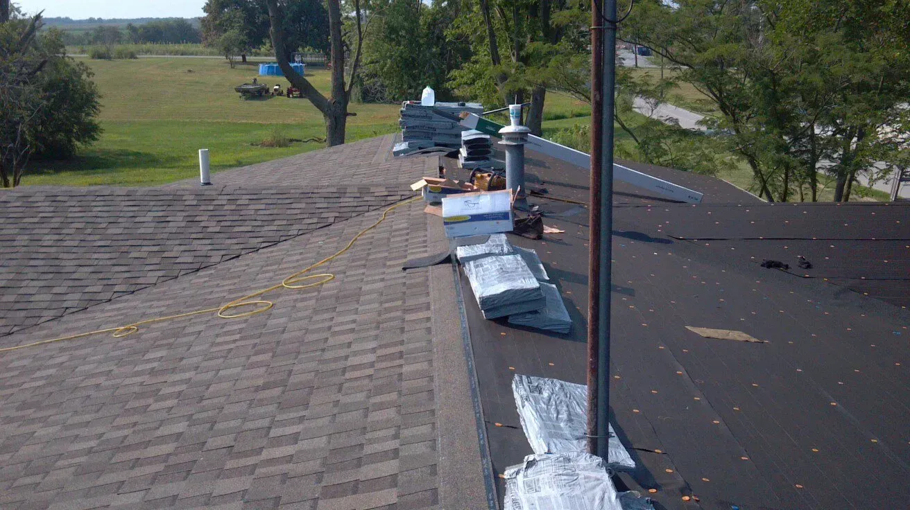 Roof under repair with workers, tools, and materials along a shingled rooftop near a grassy landscape
