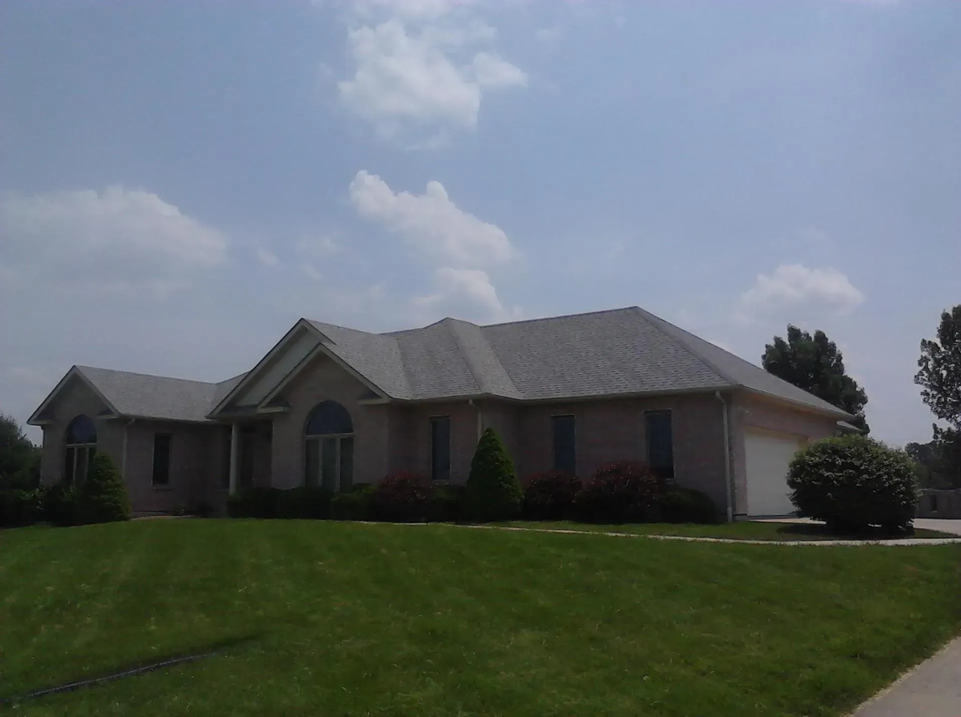 Single-story suburban house with gray roof, brick facade, and green lawn under a cloudy sky