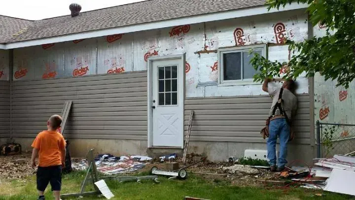 Two workers install house siding on a gray home; one stands on a ladder near a window.