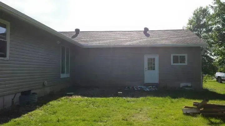 Single-story gray house with white door and small windows, shown from the backyard on a sunny day.