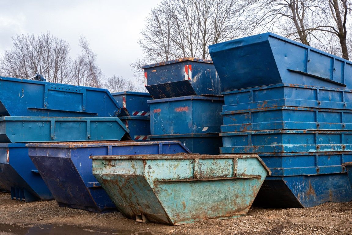 Several commercial garbage and recycling dumpsters, including a large blue bin with a red lid, lined up in a narrow urban.