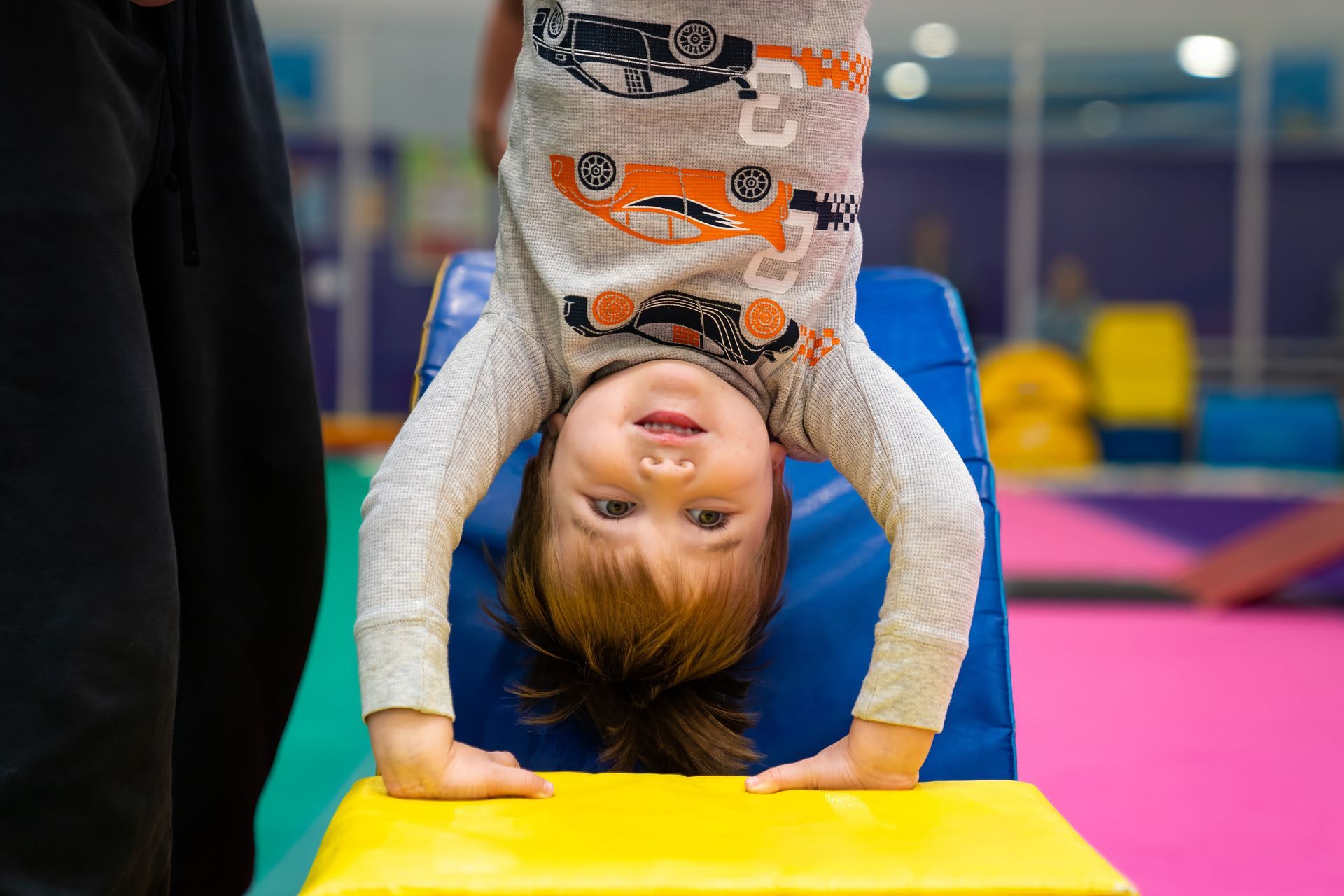 Child upside down, smiling, supported by hands on a blue and yellow gymnastics block. Indoor gym setting.