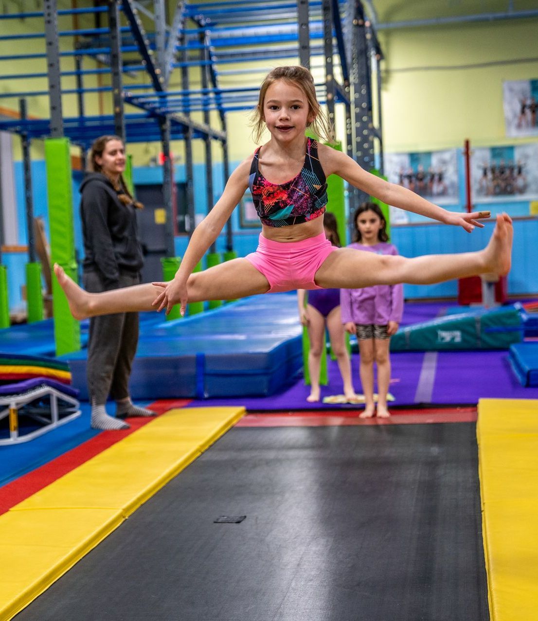 Girl in pink shorts and top does a split jump on a trampoline; other people watch in a gym.