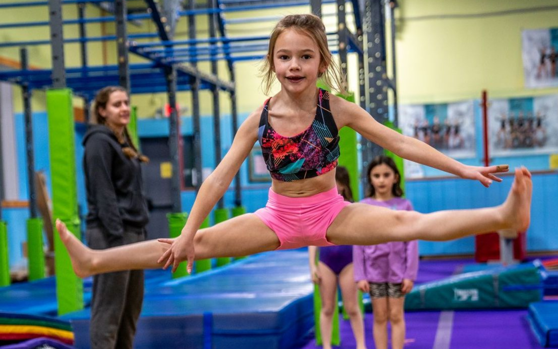 Girl in pink shorts and sports bra does the splits in a gym, two others watch.