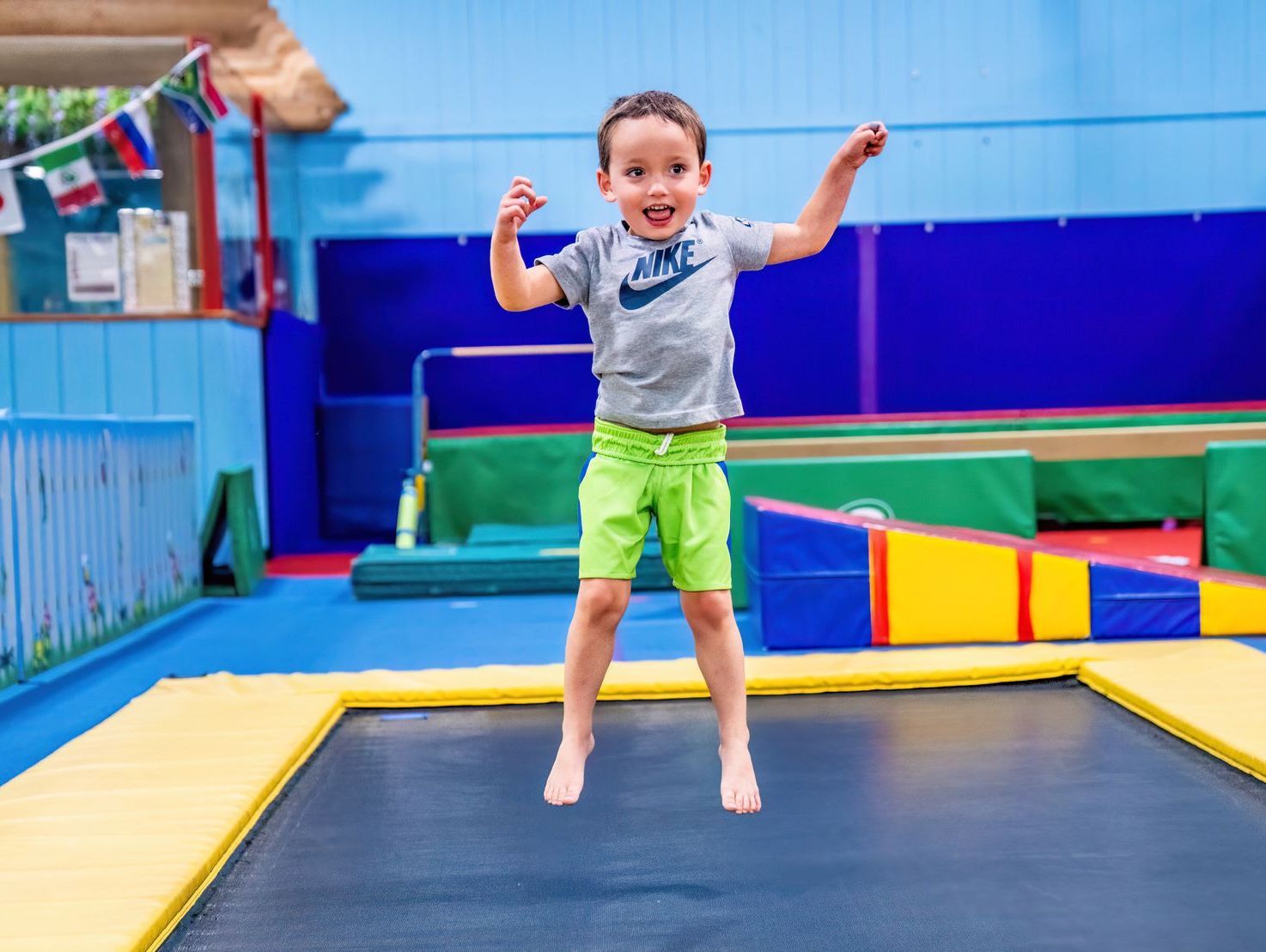 Young child jumping on a trampoline, arms raised, smiling; indoor gym setting.