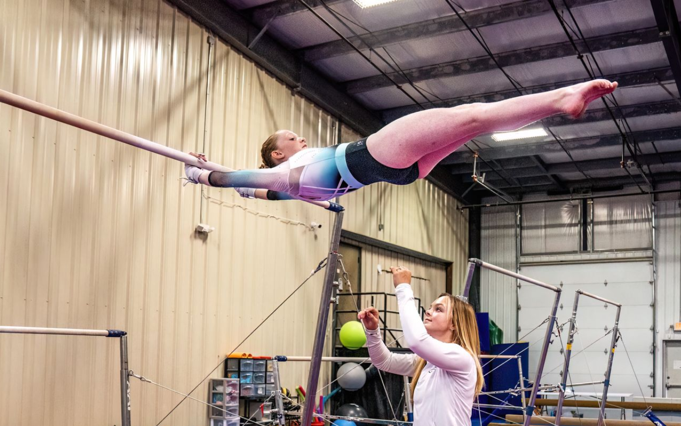 Gymnast in leotard on uneven bars, legs extended, coached by a person in a gym.
