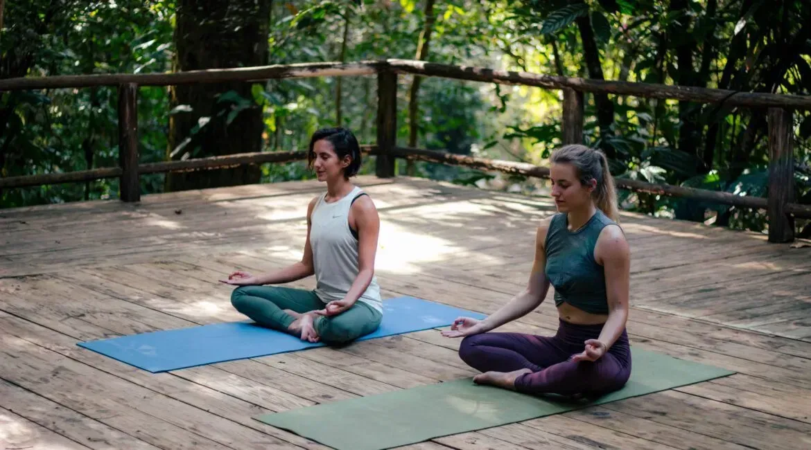 Two women are sitting on yoga mats on a wooden deck.