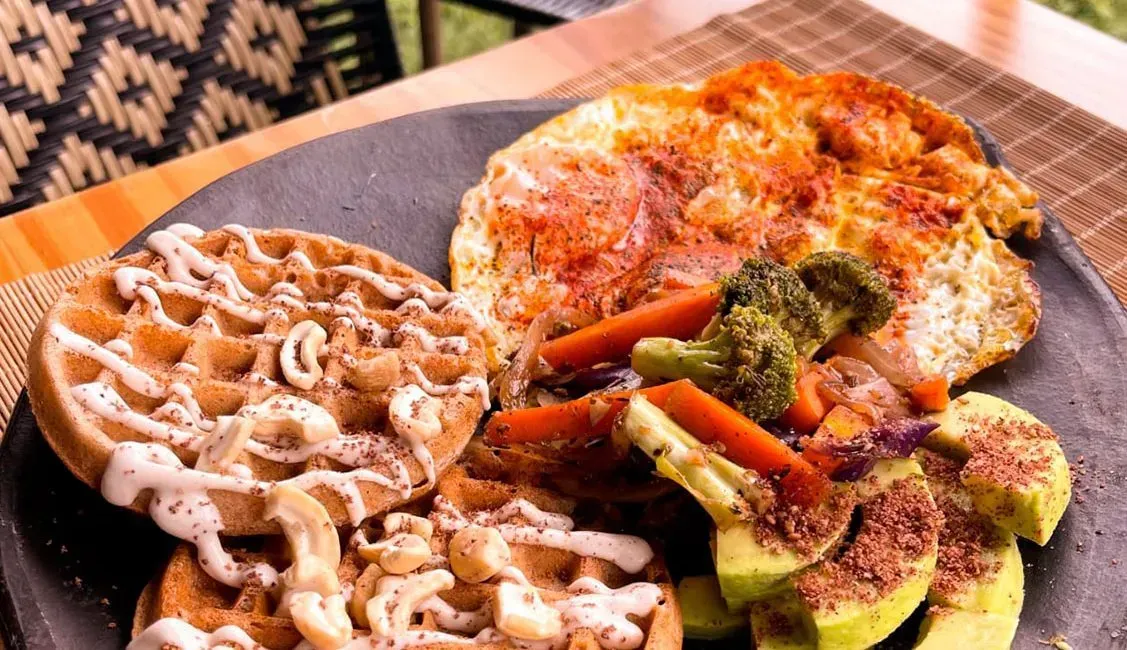 A plate of food with waffles , eggs , broccoli and avocado on a table.