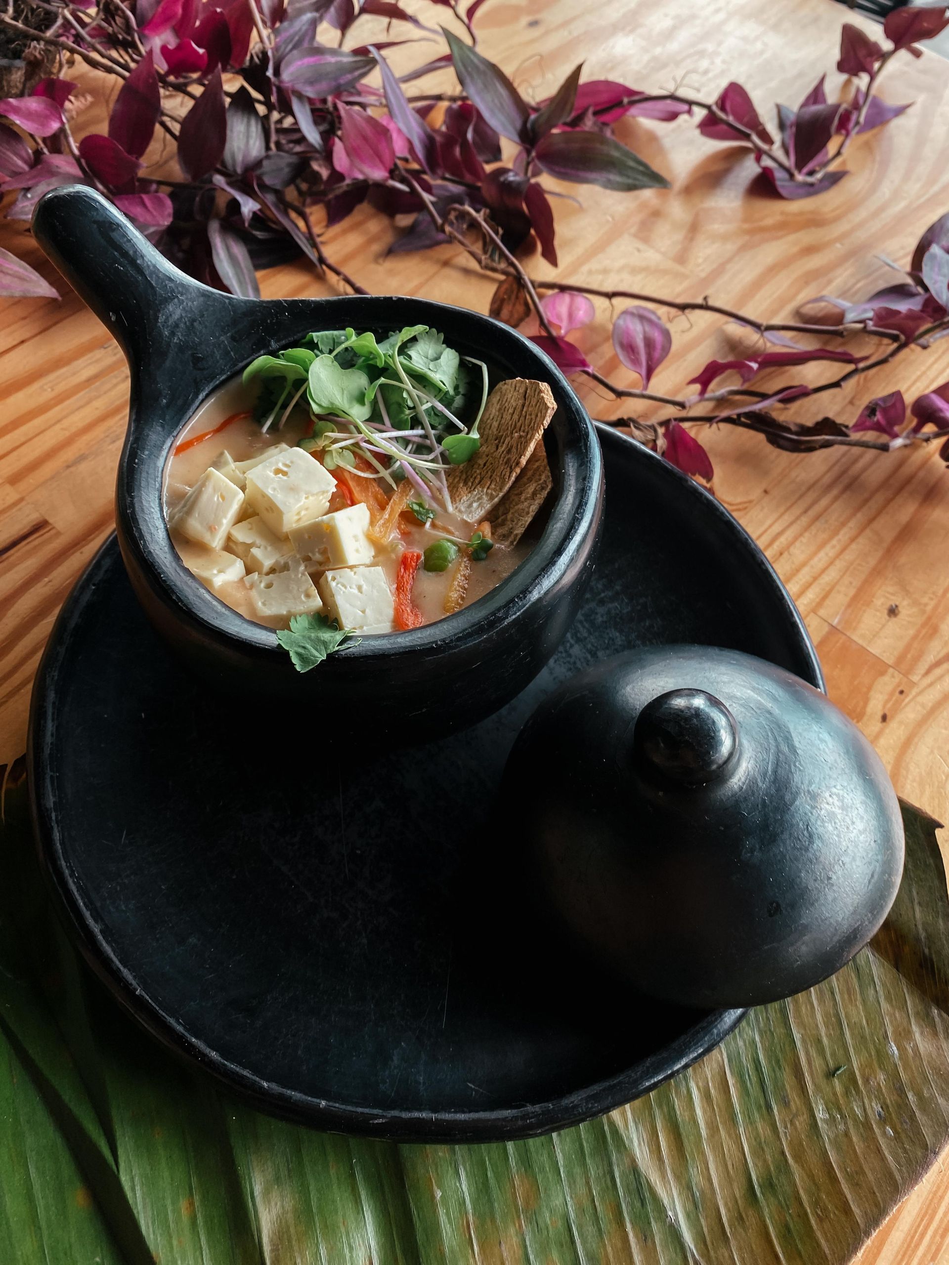 A bowl of soup is sitting on a black plate on a wooden table.