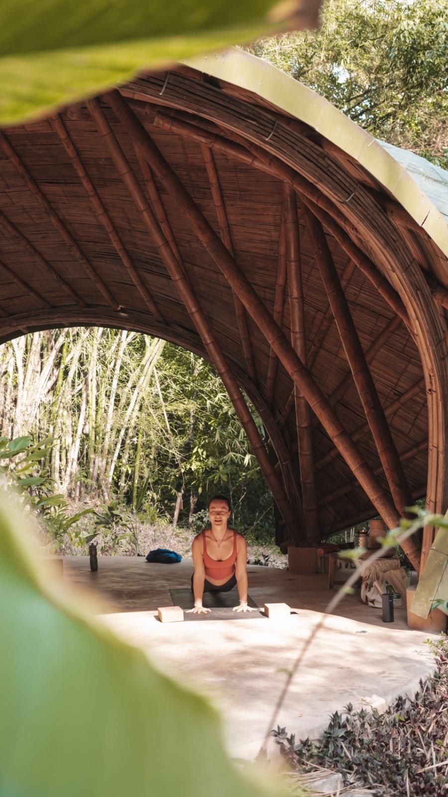A woman is sitting on a yoga mat under a wooden structure.