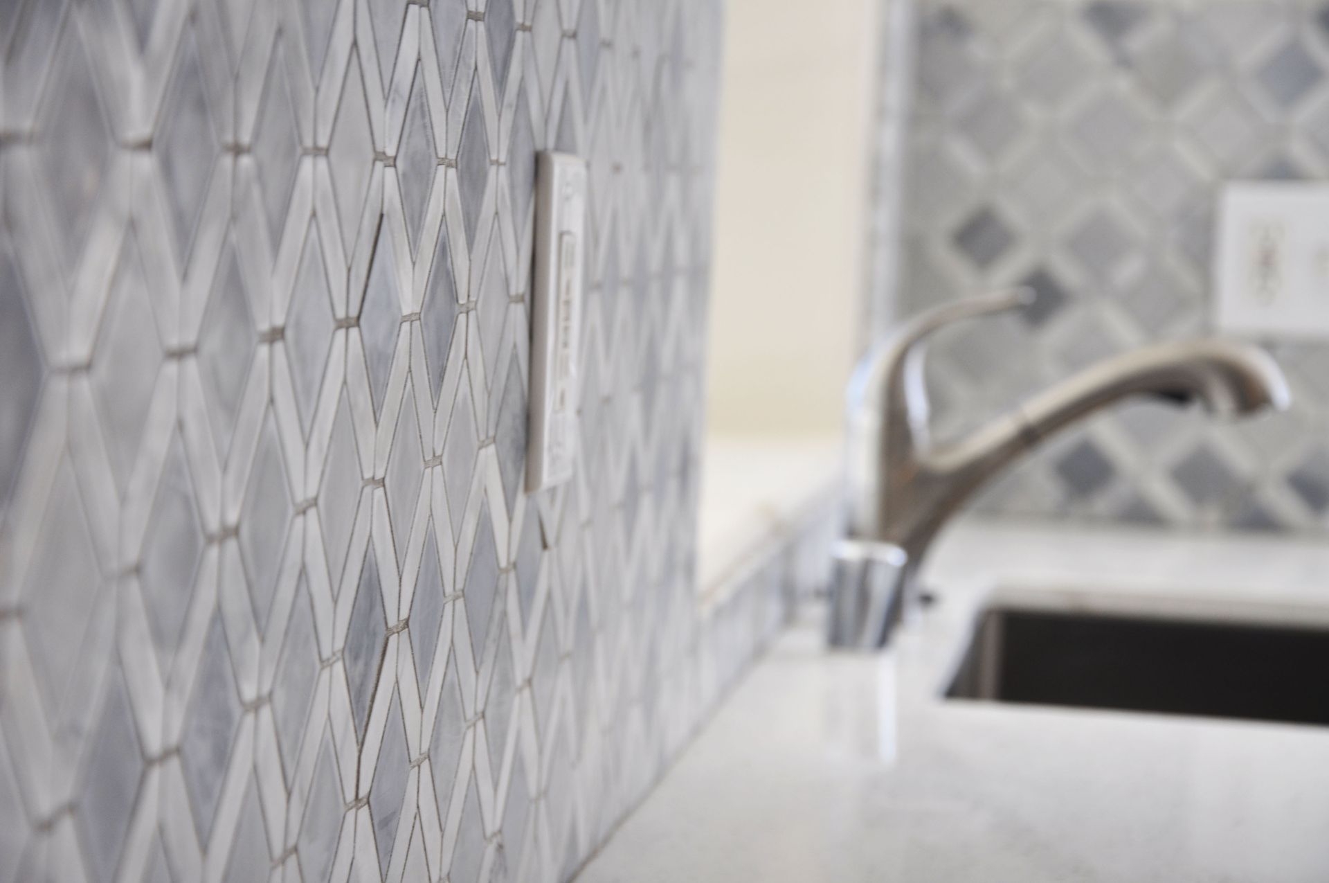 Close-up of kitchen backsplash with diamond-patterned tile in shades of gray and white; faucet and sink in background.