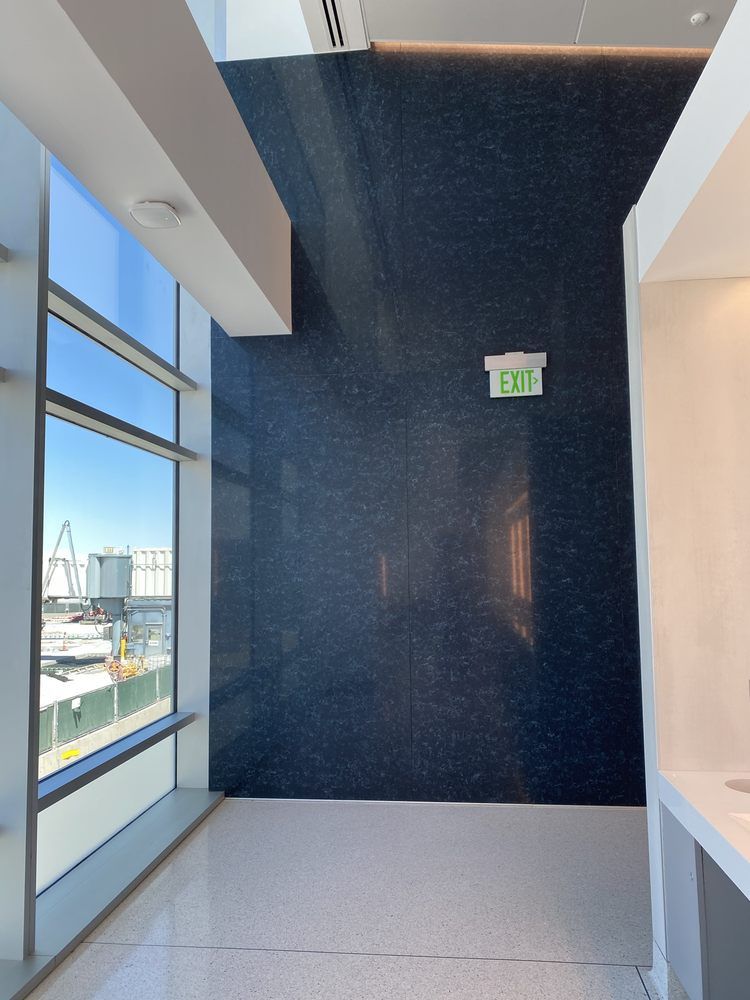 Corridor with large window, blue speckled wall, white ceiling and floor, exit sign. Airport setting.