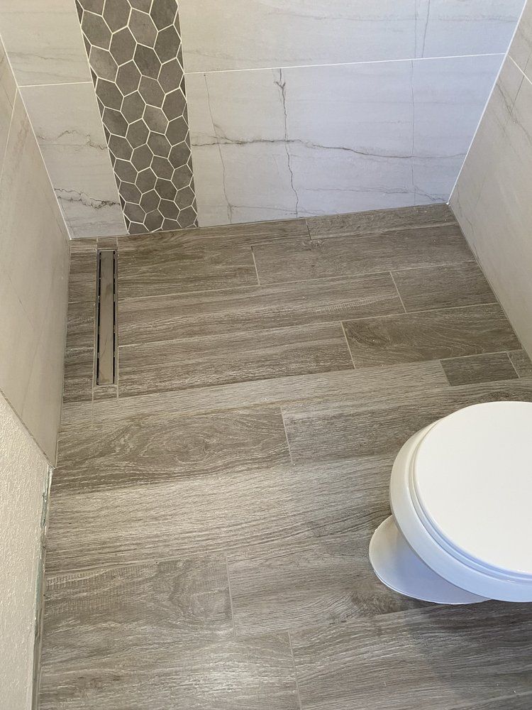 Bathroom with gray wood-look tile floor, linear drain, and white toilet.  Gray and white tiled walls.