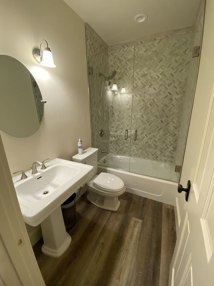 Bathroom with pedestal sink, toilet, tub/shower, and wood-look flooring. Glass shower doors.