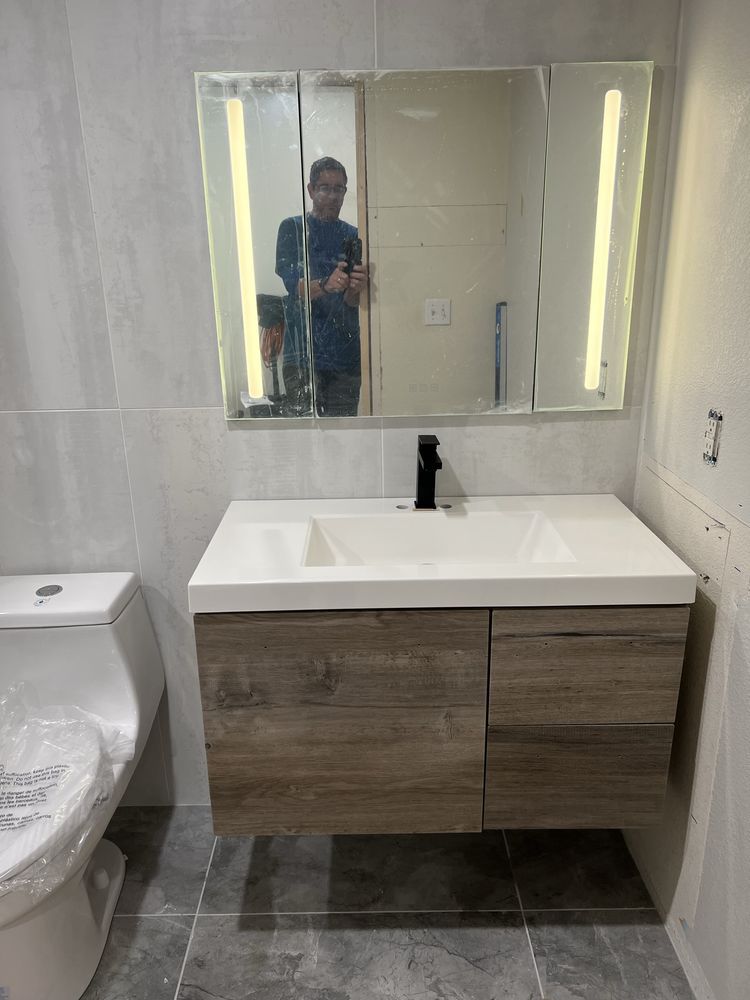 Bathroom with a white sink, wooden cabinet, and mirror with vertical lights. A person is reflected in the mirror.