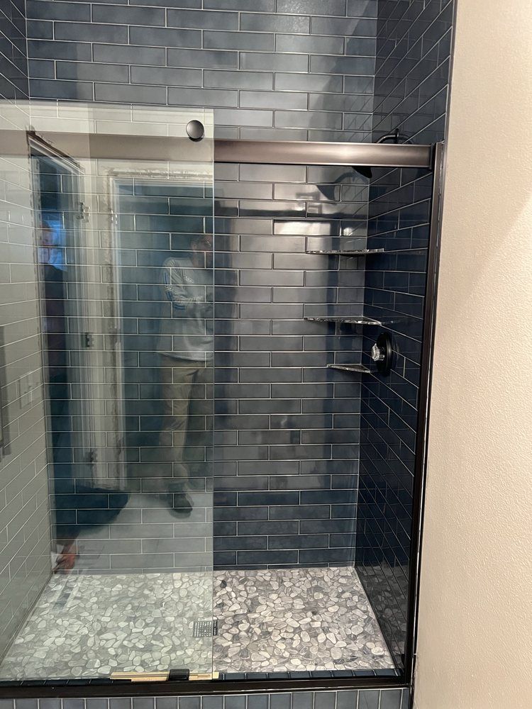 Shower with dark blue tiled walls, glass door, dark metal hardware, and mosaic floor.
