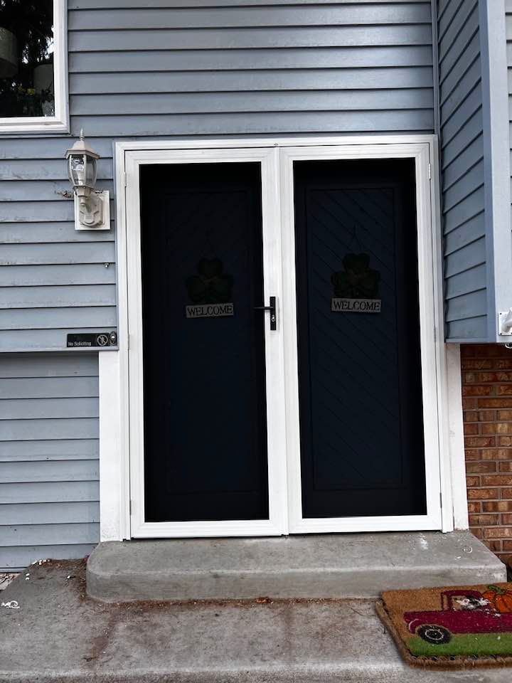 Blue house entrance with white-framed double screen doors and a welcome mat on the concrete step.