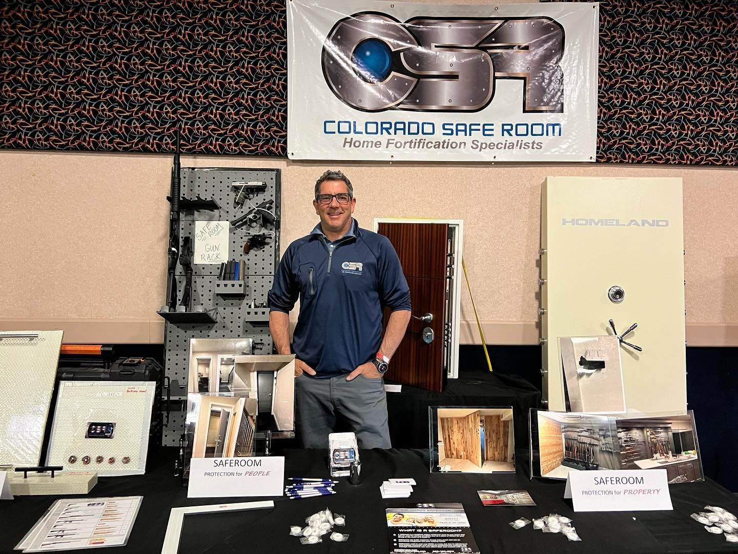 Man at a booth for Colorado Safe Room, displaying security products. Table with brochures, components, and sample doors.