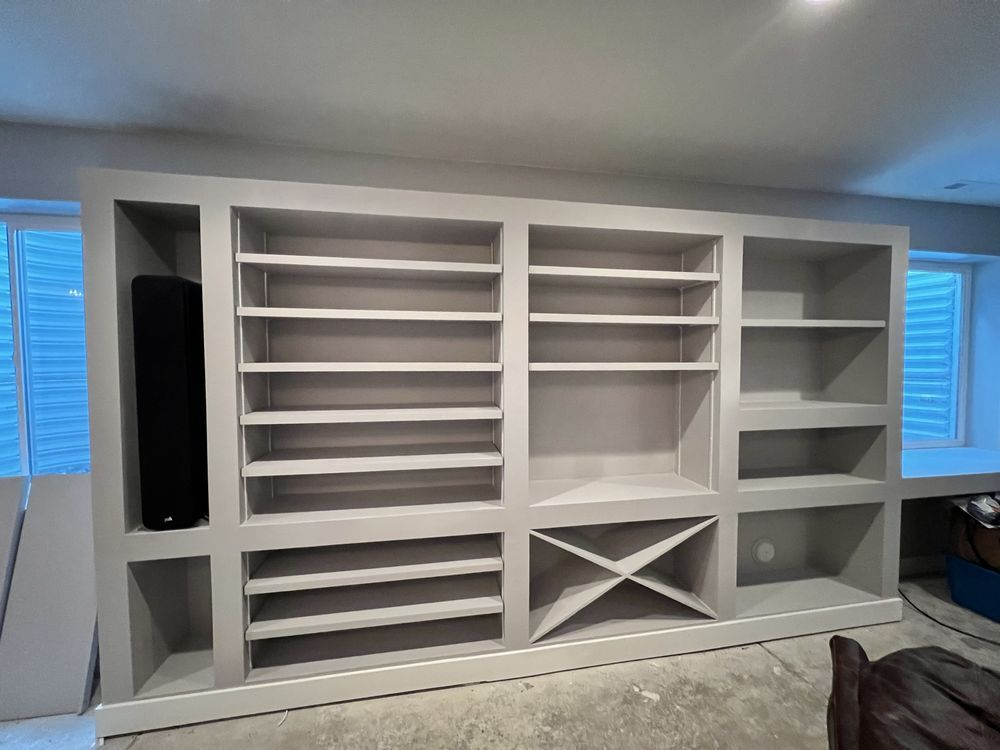 Built-in gray cabinet with shelves, wine rack, and space for a speaker in a basement room.