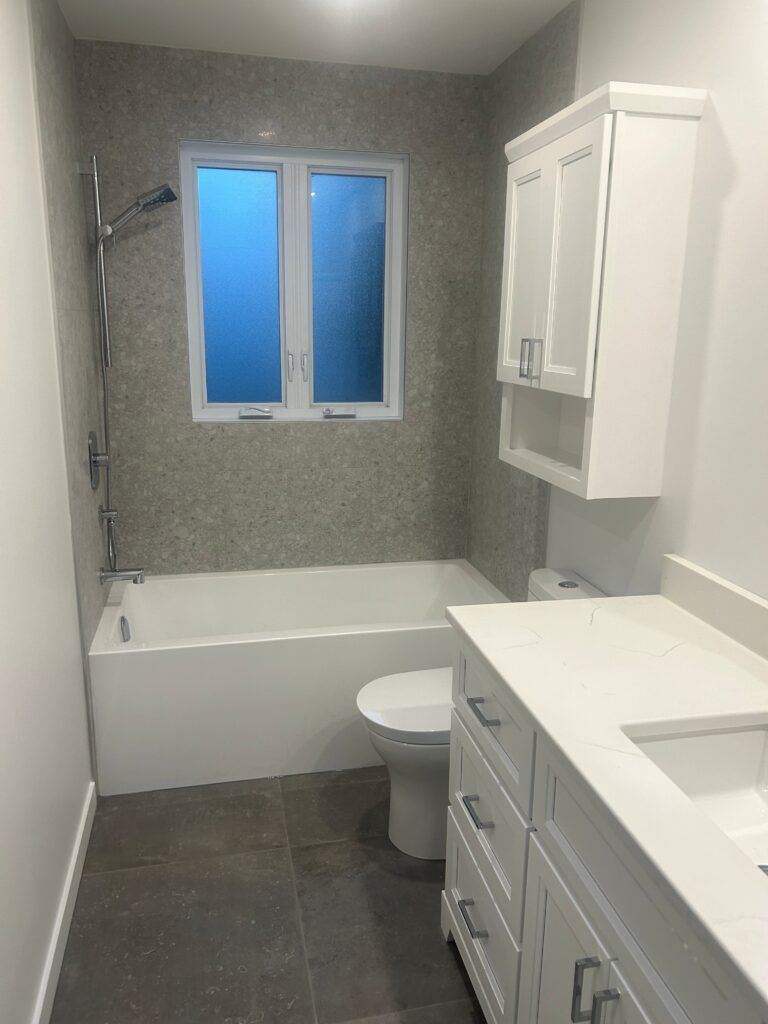 Salle de bains avec meuble-lavabo blanc, toilettes, baignoire et carrelage gris. Fenêtre en verre dépoli.