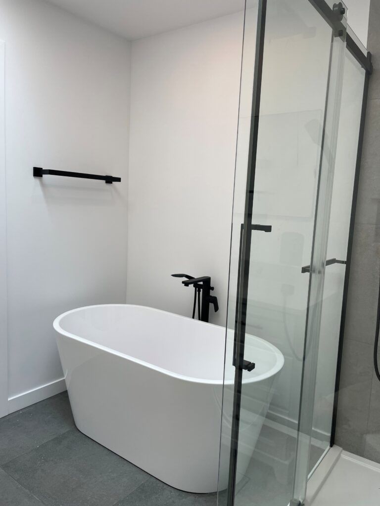 Salle de bains moderne blanche avec baignoire îlot, robinetterie noire et douche en verre.