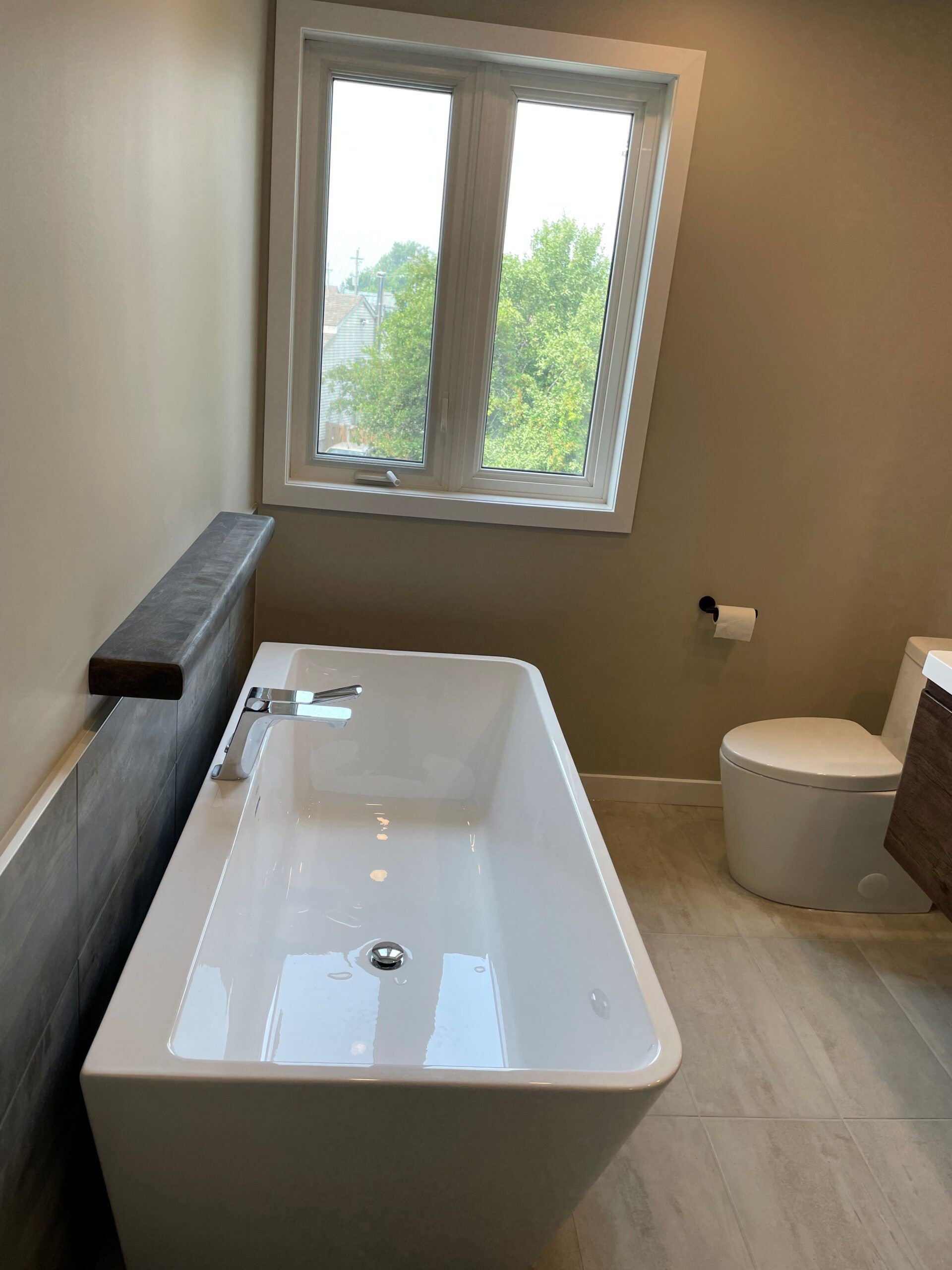 Salle de bains moderne avec baignoire rectangulaire blanche, toilettes et fenêtre. Murs aux tons neutres et touches de bois.