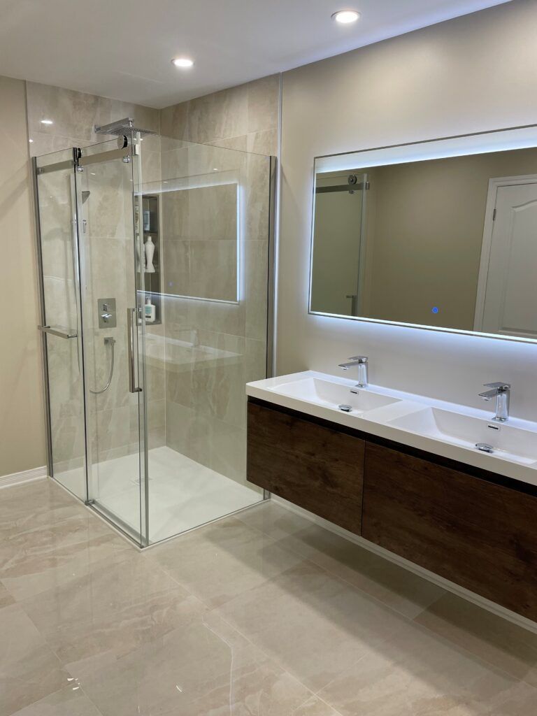 Salle de bains moderne avec douche en verre, double vasque avec miroir éclairé et sol carrelé clair.