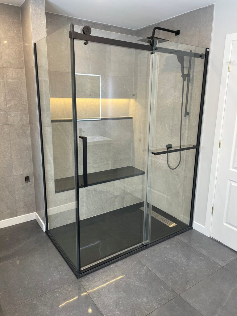 Cabine de douche moderne avec cadre noir et portes en verre transparent, sol gris foncé, banc intégré et pommeau de douche.
