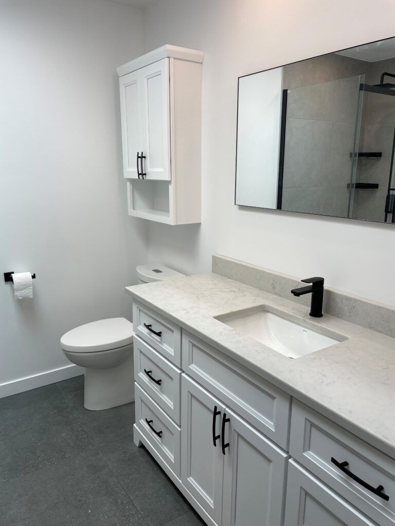 Salle de bains blanche avec meuble-lavabo, miroir, toilettes et armoire. Touches noires, sol gris.
