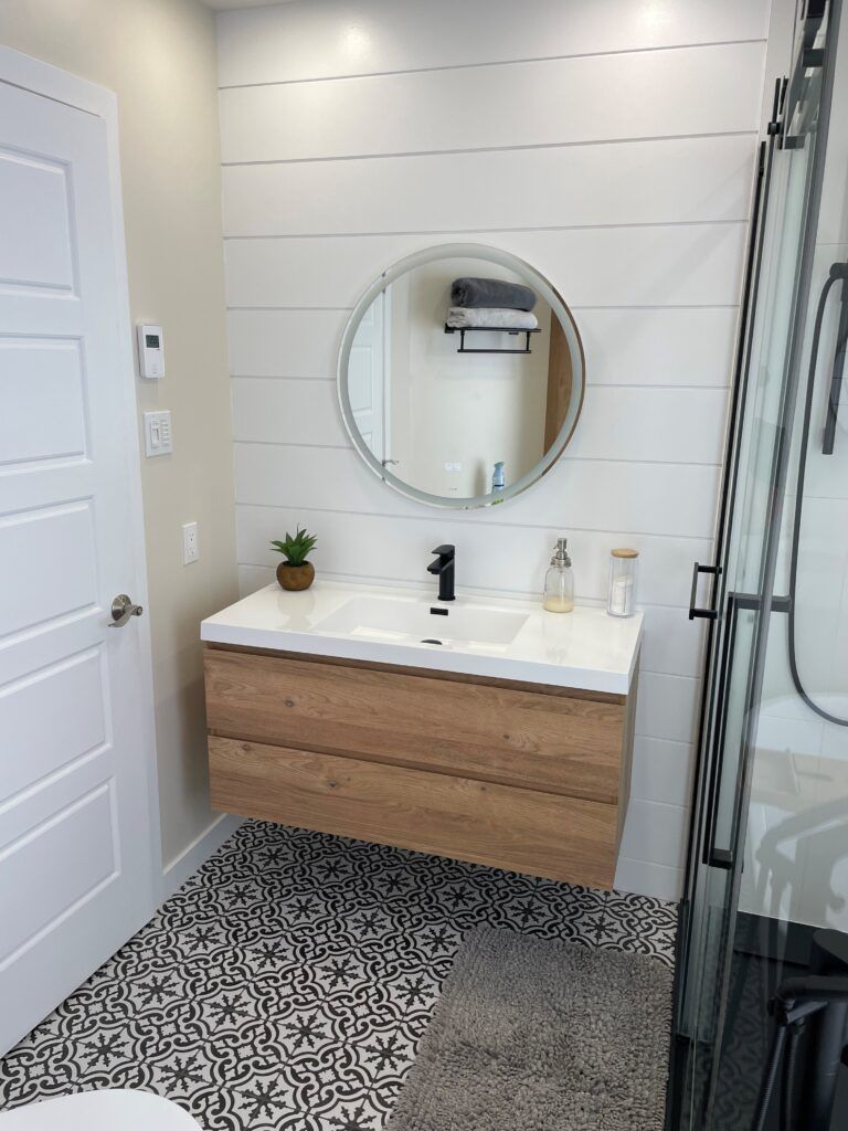 Salle de bains moderne avec meuble-lavabo suspendu en bois, miroir rond, sol à motifs et murs lambrissés blancs.