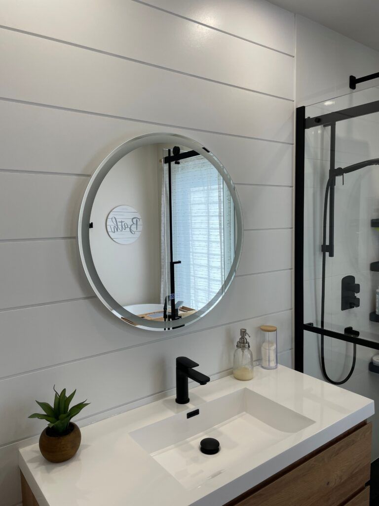 Salle de bains avec mur lambrissé blanc, miroir rond, robinet noir et cabine de douche.