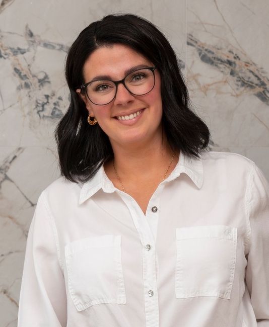 Une femme à lunettes sourit, vêtue d'une chemise blanche, devant un mur de marbre.