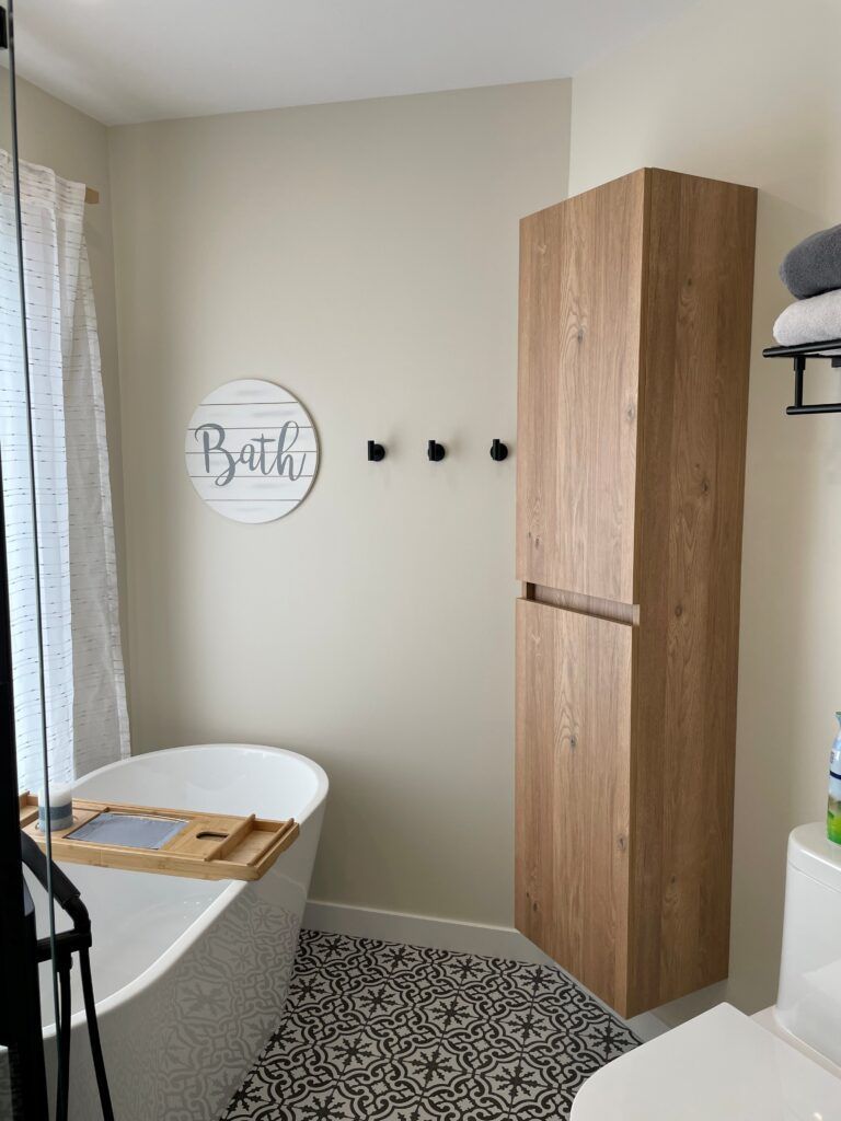Salle de bains avec baignoire blanche, haute armoire de rangement en bois et carrelage noir à motifs au sol.