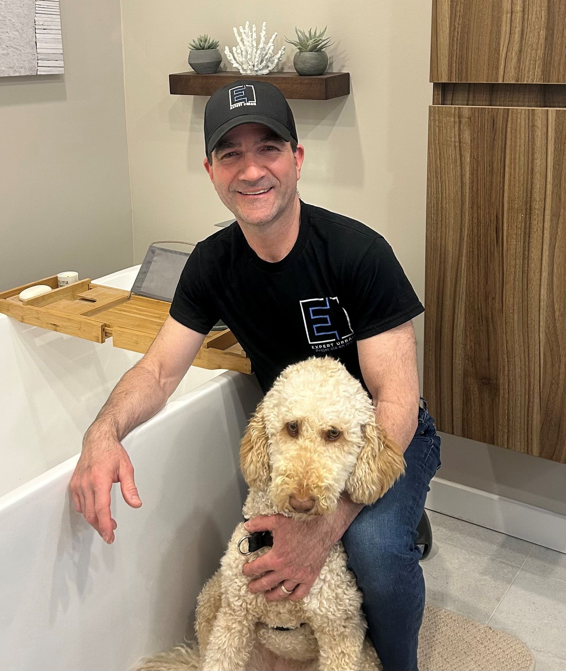 Un homme en chemise et casquette noires sourit, tenant un golden doodle. Intérieur.